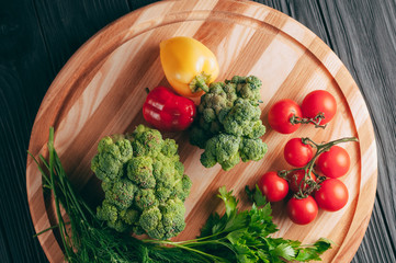 Fresh vegetables: cherry tomatoes, broccoli, pepper, dill, parsley on a round wooden board, on a dark wooden table. Recipe. Ingredients. Dietary food. Place under the text. View from above.