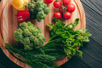 Fresh vegetables: cherry tomatoes, broccoli, pepper, dill, parsley on a round wooden board, on a dark wooden table. Recipe. Ingredients. Dietary food. Place under the text. View from above.