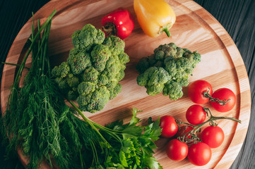 Fresh vegetables: cherry tomatoes, broccoli, pepper, dill, parsley on a round wooden board, on a dark wooden table. Recipe. Ingredients. Dietary food. Place under the text. View from above.