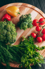 Fresh vegetables: cherry tomatoes, broccoli, pepper, dill, parsley on a round wooden board, on a dark wooden table. Recipe. Ingredients. Dietary food. Place under the text. View from above.