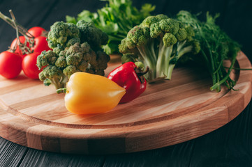 Fresh vegetables: cherry tomatoes, broccoli, pepper, dill, parsley on a round wooden board, on a dark wooden table. Recipe. Ingredients. Dietary food. Place under the text. View from above.