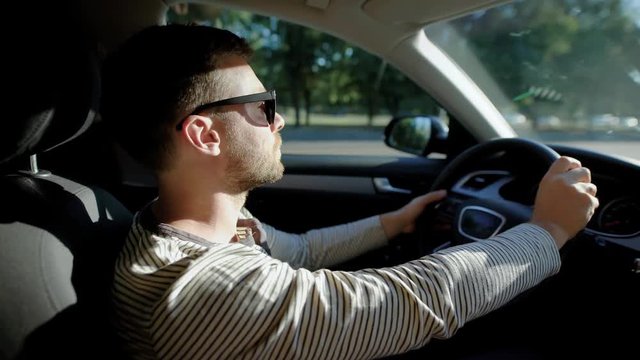 A man is driving a modern car. A handsome man with a beard in the sun protective glasses, confidently steers.
