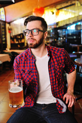 Cute young man in a plaid shirt drinking beer sitting on a chair in a pub