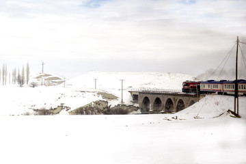 Red passenger diesel train moving on historical bridge. Snow covered railway tracks - East express between Ankara and Kars - Turkey