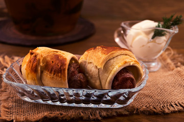 Baked meat sausages in puff pastry with white sour cream sauce on a linen napkin on a wooden background.