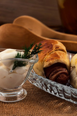 Sausage in dough, baked in the oven until golden  brown, stacked in a glass plate on a linen cloth napkin, gravy boat with sour cream and a sprig of dill, all on a wooden background.