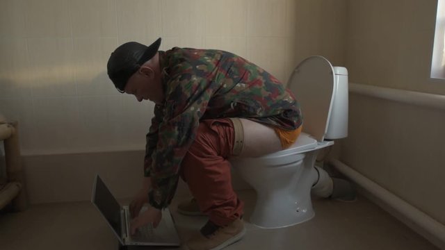 Man with gray hair, beard and mustache in military jacket with laptop sitting on toilet. Man's eye is covered with black medical patch. Dude is infatuated with computer and actively expressed emotions