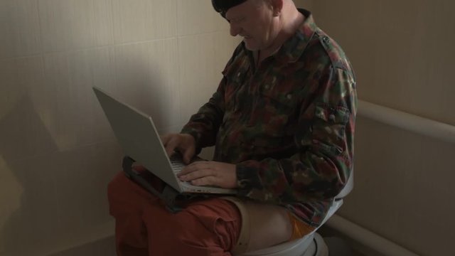Man with gray hair, beard and mustache in military jacket with laptop sitting on toilet. Man's eye is covered with black medical patch. Dude is infatuated with computer and actively expressed emotions