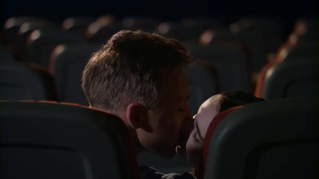 Young people kiss passionately at empty cinema hall. Love couple alone in empty cinema hall. Love couple kissing in cinema. Romantic date in movie theater in slow motion