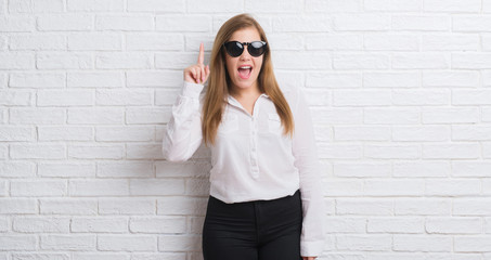 Young adult business woman standing over white brick wall wearing sunglasses surprised with an idea or question pointing finger with happy face, number one