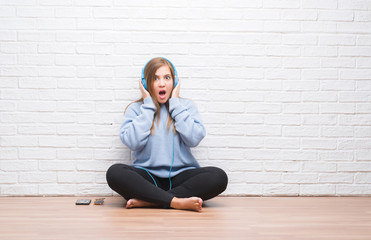 Young adult woman over white brick wall listening music wearing headphones scared in shock with a surprise face, afraid and excited with fear expression