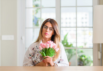 Young woman at home holding pink tulips flowers with a confident expression on smart face thinking serious