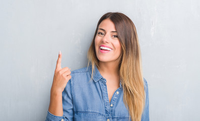 Young adult woman over grey grunge wall wearing denim outfit surprised with an idea or question pointing finger with happy face, number one