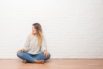Young adult woman sitting on the floor over white brick wall at home looking away to side with...