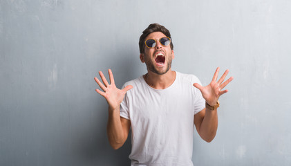 Naklejka premium Handsome young man over grey grunge wall wearing sunglasses celebrating crazy and amazed for success with arms raised and open eyes screaming excited. Winner concept