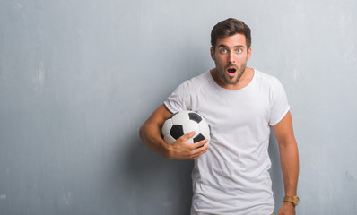 Handsome young man over grey grunge wall holding soccer football ball scared in shock with a...