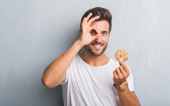 Handsome young man over grey grunge wall eating chocolate chip cooky with happy face smiling doing ok sign with hand on eye looking through fingers