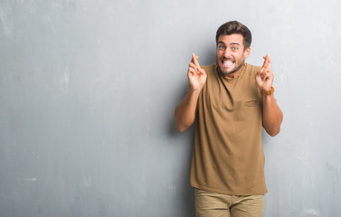 Handsome young man over grey grunge wall smiling crossing fingers with hope and eyes closed. Luck and superstitious concept.