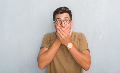 Handsome young man over grey grunge wall wearing glasses shocked covering mouth with hands for...