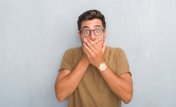 Handsome young man over grey grunge wall wearing glasses shocked covering mouth with hands for mistake. Secret concept.