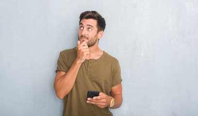 Handsome young man over grey grunge wall texting a message using smartphone serious face thinking...