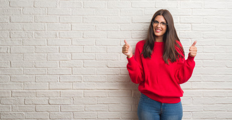 Fototapeta na wymiar Young brunette woman standing over white brick wall success sign doing positive gesture with hand, thumbs up smiling and happy. Looking at the camera with cheerful expression, winner gesture.