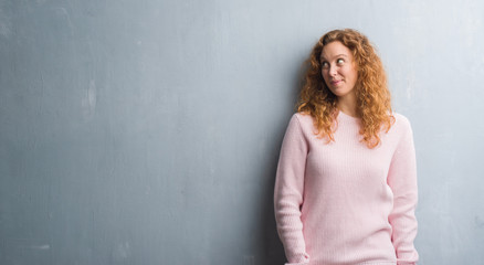 Fototapeta premium Young redhead woman over grey grunge wall wearing pink sweater smiling looking side and staring away thinking.