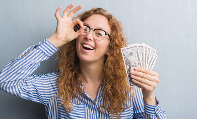 Young redhead woman over grey grunge wall holding dollars with happy face smiling doing ok sign with hand on eye looking through fingers