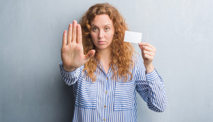 Young redhead woman over grey grunge wall holding blank visit card with open hand doing stop sign with serious and confident expression, defense gesture
