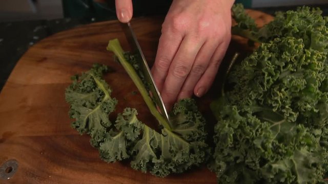 Woman in kitchen cuts kale with knife on wood chopping board