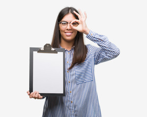 Young hispanic woman holding clipboard with happy face smiling doing ok sign with hand on eye looking through fingers