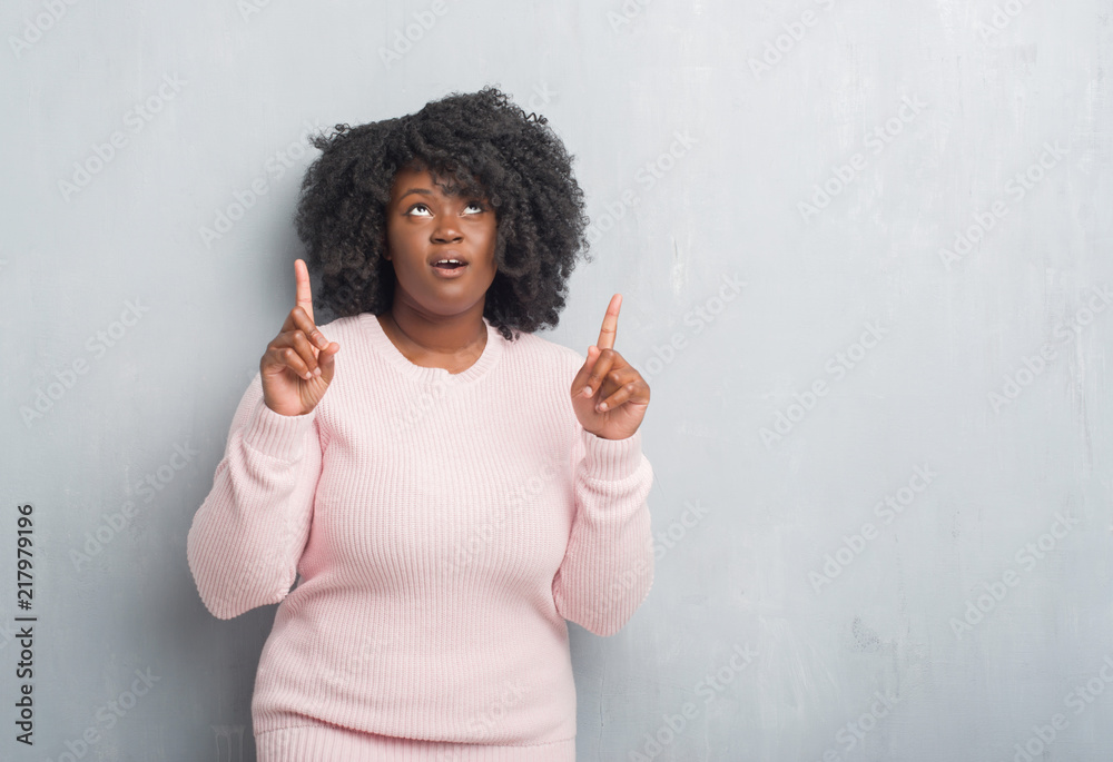 Wall mural Young african american plus size woman over grey grunge wall wearing winter sweater amazed and surprised looking up and pointing with fingers and raised arms.