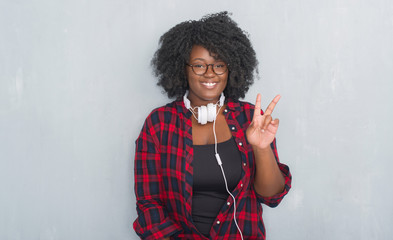 Young african american woman over grey grunge wall wearing headphones smiling with happy face winking at the camera doing victory sign. Number two.