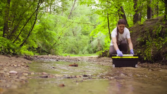 Woman scientist environmentalist sitting near the creek. She opening a tool box, taking sample of water and pouring it into the test tube.