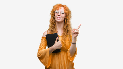 Young redhead student woman holding a book very happy pointing with hand and finger to the side