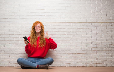 Young redhead woman sitting over brick wall talking on the phone happy with big smile doing ok sign, thumb up with fingers, excellent sign
