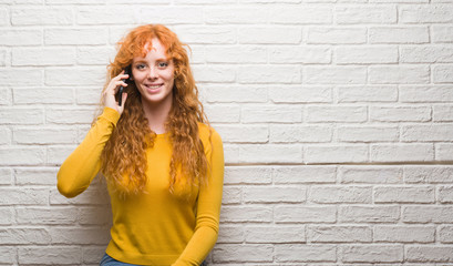 Young redhead woman standing over brick wall talking on the phone with a happy face standing and smiling with a confident smile showing teeth