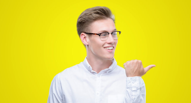 Young handsome blond man smiling with happy face looking and pointing to the side with thumb up.