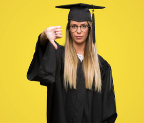 Beautiful young woman wearing glasses and graduation robe with angry face, negative sign showing dislike with thumbs down, rejection concept