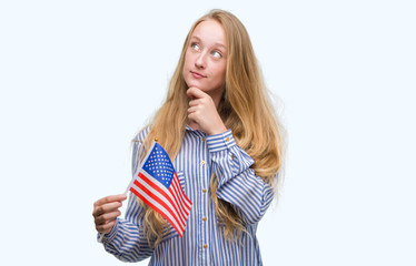 Blonde teenager woman holding flag of United States of America serious face thinking about question, very confused idea