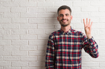 Young adult man standing over white brick wall showing and pointing up with fingers number four while smiling confident and happy.