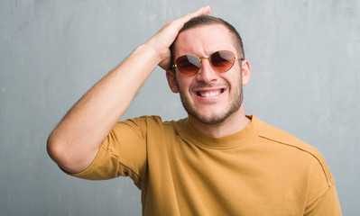 Young caucasian man over grey grunge wall wearing sunglasses stressed with hand on head, shocked with shame and surprise face, angry and frustrated. Fear and upset for mistake.