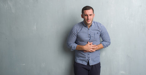 Obraz premium Young caucasian man over grey grunge wall with hand on stomach because indigestion, painful illness feeling unwell. Ache concept.