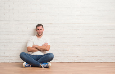 Obraz na płótnie Canvas Young caucasian man sitting on the floor over white brick wall skeptic and nervous, disapproving expression on face with crossed arms. Negative person.
