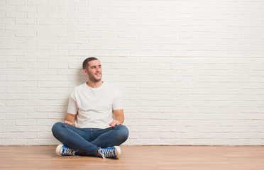 Young caucasian man sitting on the floor over white brick wall looking away to side with smile on face, natural expression. Laughing confident.