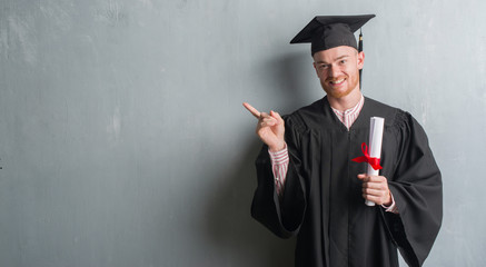 Young redhead man over grey grunge wall wearing graduate uniform holding degree very happy pointing with hand and finger to the side