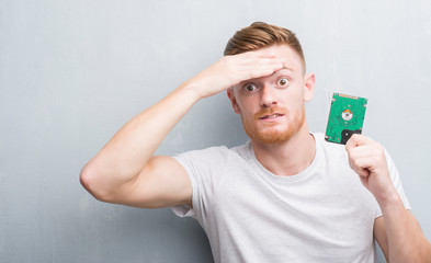 Young redhead man over grey grunge wall holding hard drive stressed with hand on head, shocked with...