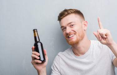 Young redhead man over grey grunge wall drinking beer bottle surprised with an idea or question pointing finger with happy face, number one