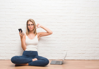 Beautiful young woman sitting on the floor texting using smartphone and laptop with angry face,...