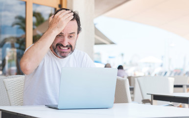 Handsome senior man using laptop at restaurant stressed with hand on head, shocked with shame and surprise face, angry and frustrated. Fear and upset for mistake.